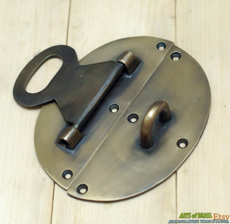 May include: A brass oval-shaped door latch with a keyhole and a hook. The latch has two hinges and is designed to be mounted on a door.