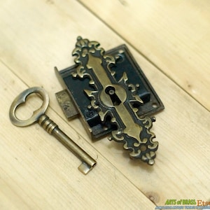May include: Antique brass door lock with a key. The lock has a decorative design with a black finish and a brass keyhole. The key is ornate and has a brass finish.