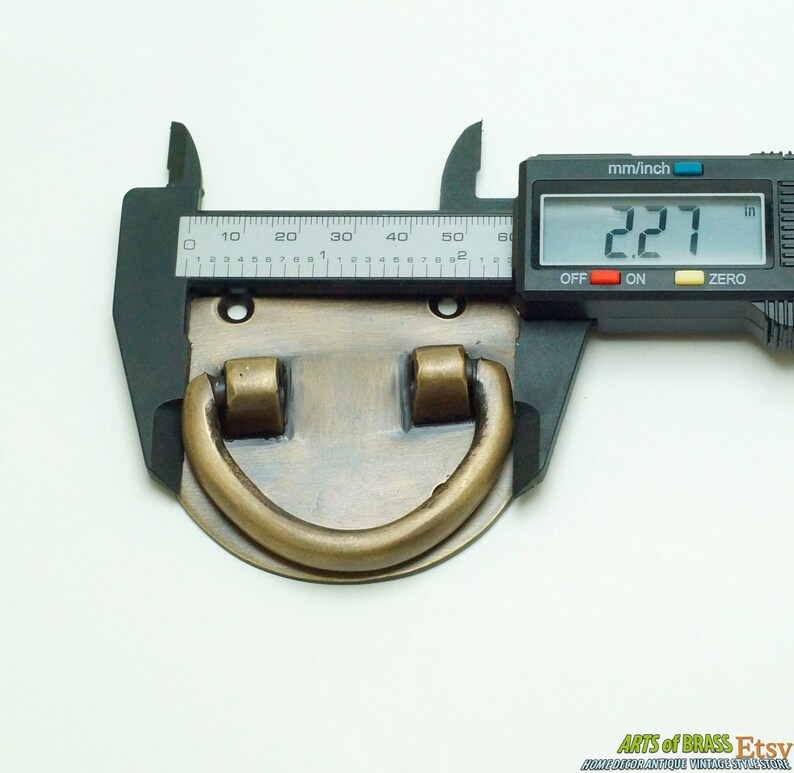 Puede incluir: Un tirador de puerta de lat&oacute;n con una base rectangular redondeada y un asa de anillo. El tirador tiene un di&aacute;metro de 5,77 cm.