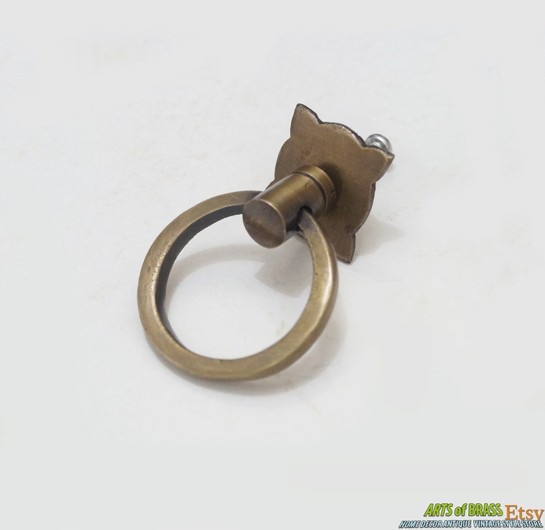 Puede incluir: Una manija de puerta de armario de anillo de lat&oacute;n con una placa posterior decorativa. La manija es una forma de anillo simple y la placa posterior tiene un dise&ntilde;o floral.