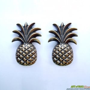 Op de afbeelding: Twee bronzen ananasvormige meubelgrepen. Elke greep heeft een gedetailleerd ananasontwerp met een getextureerde schil en een bladerkroon. De grepen zijn waarschijnlijk van metaal en geschikt voor meubels of decoratieve doeleinden.