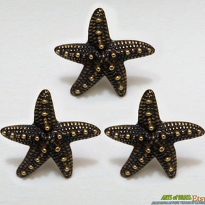 Peut inclure: Trois ornements en forme d'étoile de mer en métal doré avec une surface texturée et de petits points en relief. Les étoiles de mer sont disposées en forme de triangle sur un fond blanc.