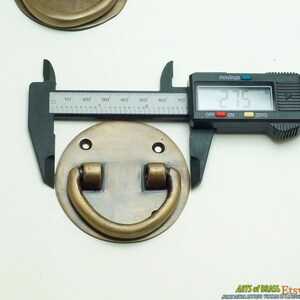 Puede incluir: Un tirador de puerta de lat&oacute;n con una base redonda y un asa en forma de D. El tirador tiene un di&aacute;metro de 5,46 cm.