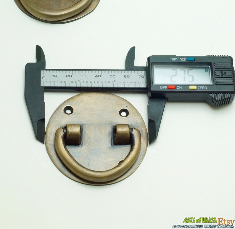 Puede incluir: Un tirador de puerta de lat&oacute;n con una base redonda y un asa en forma de D. El tirador tiene un di&aacute;metro de 5,46 cm.