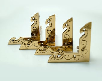 May include: Four ornate brass corner brackets with a decorative floral design. The brackets are arranged in a row, with the largest bracket at the bottom and the smallest bracket at the top.
