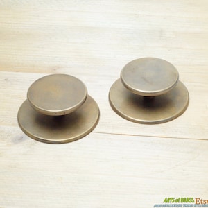Pode incluir: Dois botões de porta de latão antigo com base redonda e topo redondo. Os botões têm um acabamento desgastado e envelhecido.