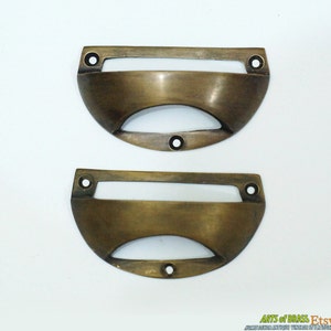 Può includere: Due tiretti per cassetti in ottone antico con un ritaglio rettangolare e un fondo curvo. I tiretti hanno una finitura usurata e invecchiata.