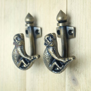 May include: Two antique brass monkey hooks with a decorative finial on top. The hooks are in the shape of a monkey with its arms wrapped around the hook.