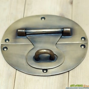 May include: A brass oval-shaped latch with a sliding bar and a ring handle. The latch has a distressed finish and is designed for use on a wooden door or chest.