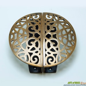 Puede incluir: Un par de tiradores de puerta de latón adornados con un diseño circular recortado. Los tiradores están separados por un pequeño espacio y tienen un acabado marrón oscuro.