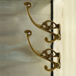 Pode incluir: Dois ganchos de latão ornamentados com um design floral decorativo. Os ganchos estão montados em uma parede branca.