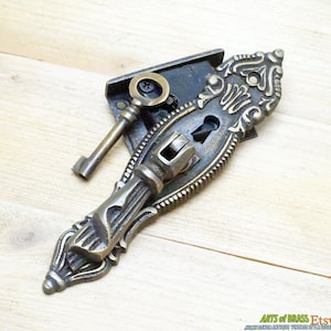 Könnte beinhalten: Antiker Türgriff aus Messing mit einem Schlüsselloch und einem dekorativen Design. Der Griff hat ein Schlüsselloch und ein dekoratives Design. Der Griff ist an einer Metallplatte mit einem Schlüsselloch befestigt. Der Griff ist aus Messing und hat eine dunkle Patina. Der Griff ist in gutem Zustand und hat einen Vintage-Look.