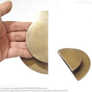 May include: Two brass half-circle door pulls, one held in a hand, the other on a white surface. The pulls are stacked on top of each other, creating a layered effect.