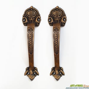 May include: A pair of antique brass door handles with a crown design and wheat stalk detailing. The handles are ornate and have a vintage look.