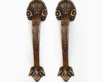May include: A pair of antique brass door handles with a crown design and wheat stalk detailing. The handles are ornate and have a vintage look.