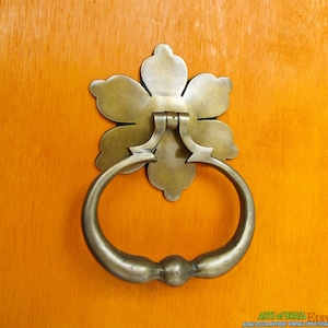May include: A brass door knocker with a flower-shaped design and a ring pull. The knocker is mounted on a wooden door.