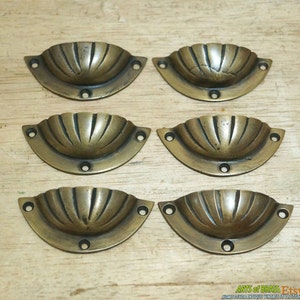 Może przedstawiać: Sześć antycznych mosiężnych gałek do szuflad w kształcie kubka z żebrowanym wzorem. Gałki mają lekko postarzaną patynę i są ułożone w dwóch rzędach po trzy.