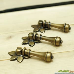 Op de afbeelding: Drie antieke messing ladegrepen met een bloemmotief. Elke greep heeft een ronde knop en een lange, slanke handgreep.