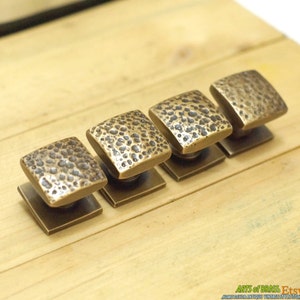 May include: Four square brass cabinet knobs with a hammered texture. The knobs are arranged in a row on a wooden surface.
