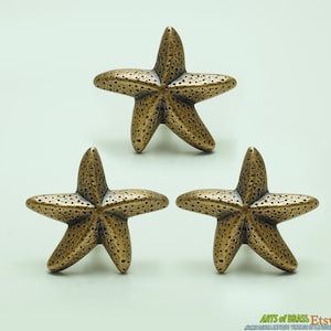 Op de afbeelding: Drie gouden metalen stervormige ladegrepen. De zeesterren hebben een getextureerd oppervlak en zijn ongeveer 5 cm in diameter.