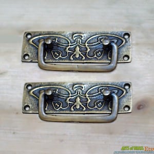 Könnte beinhalten: Zwei antike Messing-Schubladengriffe mit einem Schmetterlingsdesign. Die Griffe sind rechteckig mit einem erhabenen Schmetterlingsdesign und einem gebogenen Griff.