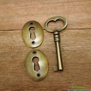 May include: Two antique brass keyhole escutcheons and a matching key. The escutcheons are oval shaped with a keyhole in the center. The key has a decorative handle and a straight shaft.