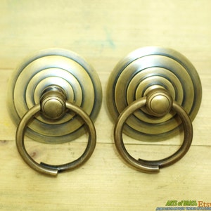 Puede incluir: Dos tiradores de puerta de armario de latón antiguo con un diseño circular y un asa de anillo. Los tiradores tienen un acabado desgastado y un patrón de círculos concéntricos.