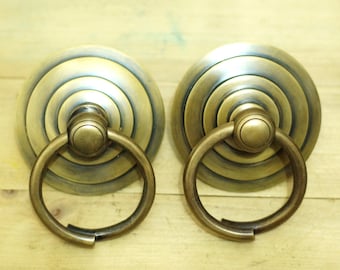 May include: Two antique brass cabinet door pulls with a circular design and a ring handle. The pulls have a distressed finish and a concentric circle pattern.