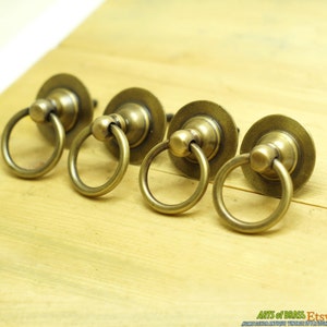May include: Four antique brass ring cabinet pulls with a round base. The pulls have a smooth, polished finish.