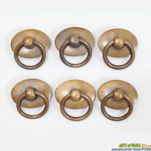 May include: Six antique brass oval cabinet drawer pulls with ring handles. Each pull has a raised round knob in the center.