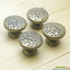 Puede incluir: Cuatro pomos de armario de latón antiguo con acabado martillado. Los pomos son redondos con un borde elevado y una parte superior lisa ligeramente abovedada.