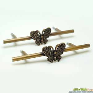 May include: Two antique brass cabinet pulls. Each pull features a butterfly design in the centre of a rectangular bar. The butterfly has detailed wing patterns. The pulls are mounted on a white background.