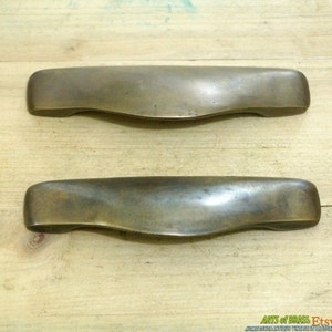 May include: Two antique brass cabinet door pulls with a curved, slightly rounded shape. The pulls are made of solid brass and have a slightly aged, tarnished finish.