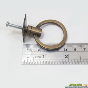 Puede incluir: Una manija de anillo de tracci&oacute;n de lat&oacute;n con una placa posterior cuadrada y un tornillo. La manija tiene un di&aacute;metro de 5 cm. La manija se mide con una regla.