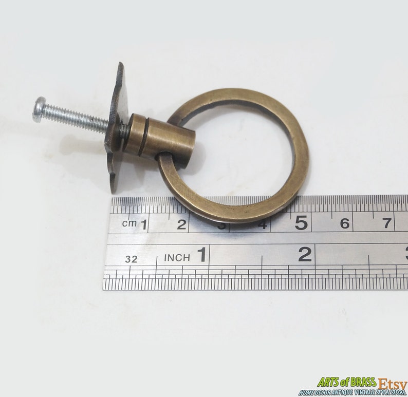 Puede incluir: Una manija de anillo de tracci&oacute;n de lat&oacute;n con una placa posterior cuadrada y un tornillo. La manija tiene un di&aacute;metro de 5 cm. La manija se mide con una regla.