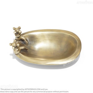 May include: A small, oval-shaped brass soap dish with two decorative faucets on the side. The dish is made of polished brass and has a smooth, shiny finish.