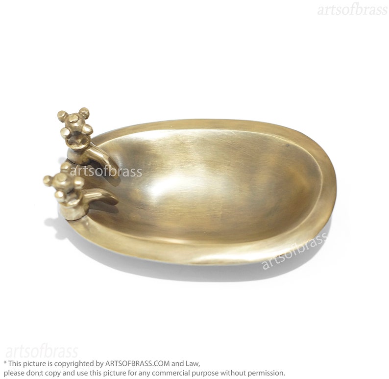 May include: A small, oval-shaped brass soap dish with two decorative faucets on the side. The dish is made of polished brass and has a smooth, shiny finish.