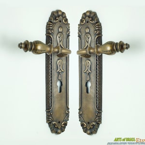 May include: A pair of antique brass door handles with ornate designs. The handles are rectangular with a keyhole and a decorative knob. The handles are in a distressed finish.