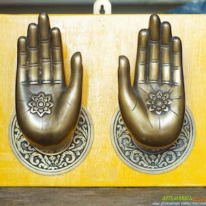 May include: A pair of ornate brass hand-shaped door handles with intricate floral designs on a yellow wooden background.