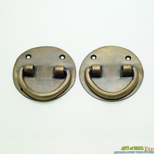 Pode incluir: Duas puxadores de gaveta de latão antigo com uma alça redonda em forma de anel. As puxadores são circulares e têm um acabamento liso e envelhecido.