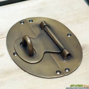 May include: A brass oval-shaped latch with a hasp and a sliding bolt. The latch has a smooth, polished finish and is designed for use on a wooden chest or box.