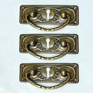 May include: Three antique brass drawer pulls with ornate designs. Each pull features a keyhole, a decorative wreath, and a curved handle.