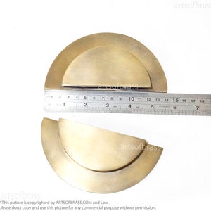 May include: Two brass half-circle cabinet door pulls, one slightly larger than the other, are shown on a white surface. A ruler is placed below the pulls to show their size.