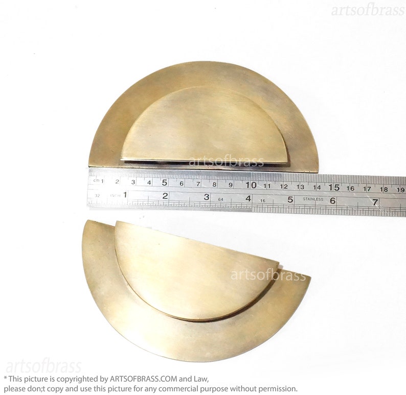 May include: Two brass half-circle cabinet door pulls, one slightly larger than the other, are shown on a white surface. A ruler is placed below the pulls to show their size.