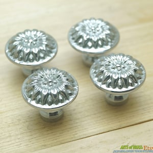 May include: Four silver-colored cabinet knobs with a floral design. Each knob features a rounded, textured top with a detailed flower pattern and a short, cylindrical base. The knobs are arranged on a light-colored wooden surface.