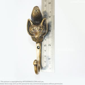 May include: A brass fox hook with a detailed design. The hook is about 3 inches long and has a rounded top. The fox's face is facing forward with a black nose and eyes.