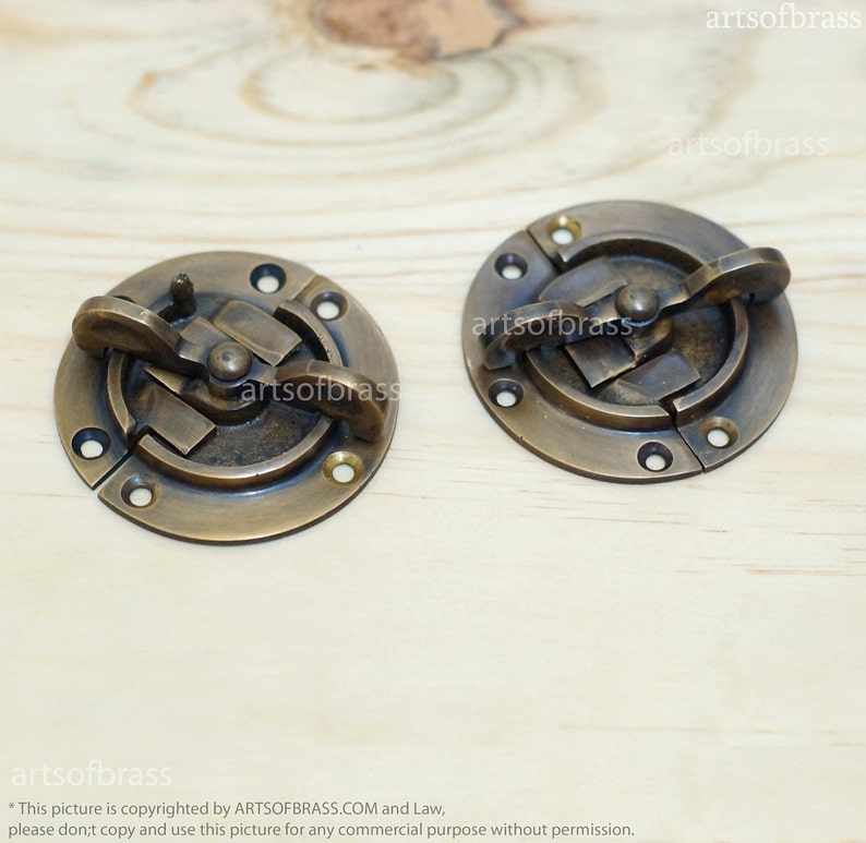 May include: Two antique brass cabinet door catches with a round shape and a latch mechanism. The catches are on a wooden surface.