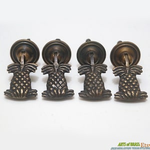 Pode incluir: Conjunto de quatro puxadores de gaveta de abacaxi de latão antigo. Cada puxador apresenta um design de abacaxi detalhado com uma base redonda.
