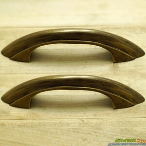 May include: Two antique brass cabinet pulls with a curved design. The pulls have a distressed finish and are approximately 7.6 cm long.