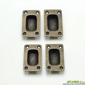 May include: Four antique brass cabinet pulls with a rectangular shape and a recessed centre. The pulls have four small holes for mounting.